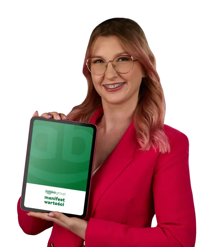 Uśmiechnięta kobieta w czerwonej marynarce trzyma w dłoni zieloną książeczkę zatytułowaną Culture Book Żabka.